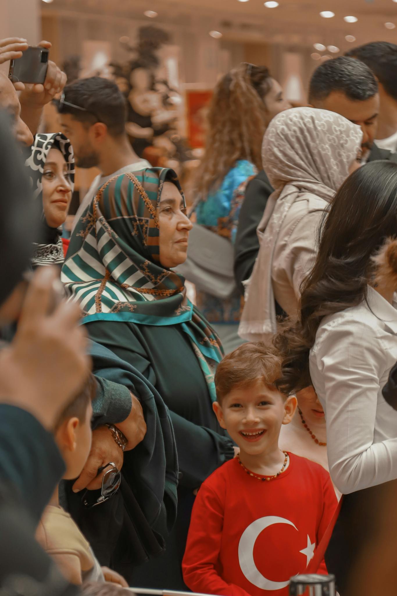 A joyful boy in a red Turkish shirt amidst a diverse crowd at a cultural gathering.